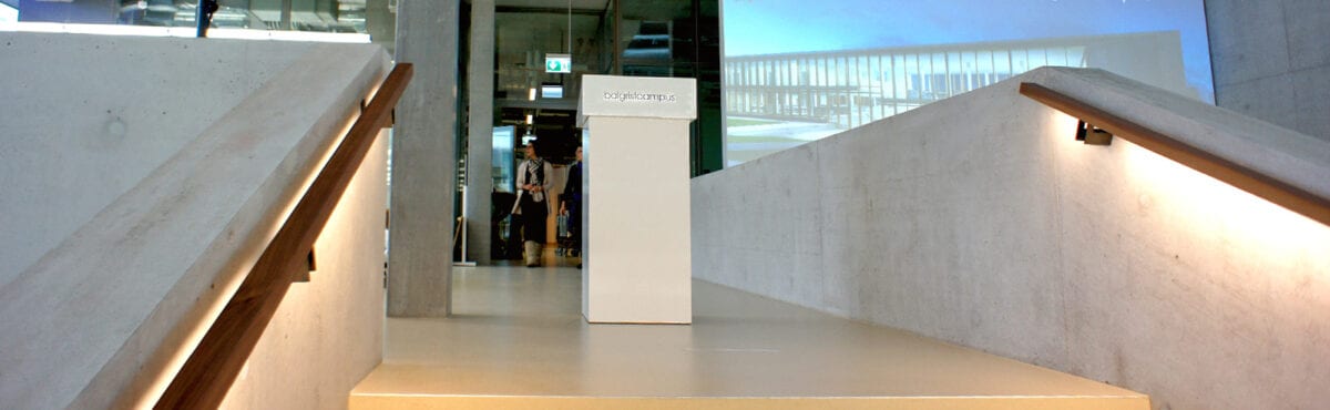 Moderner Eingangsbereich mit Sichtbetonwand, beleuchtetem Handlauf aus Holz und Blick auf eine Glasfront