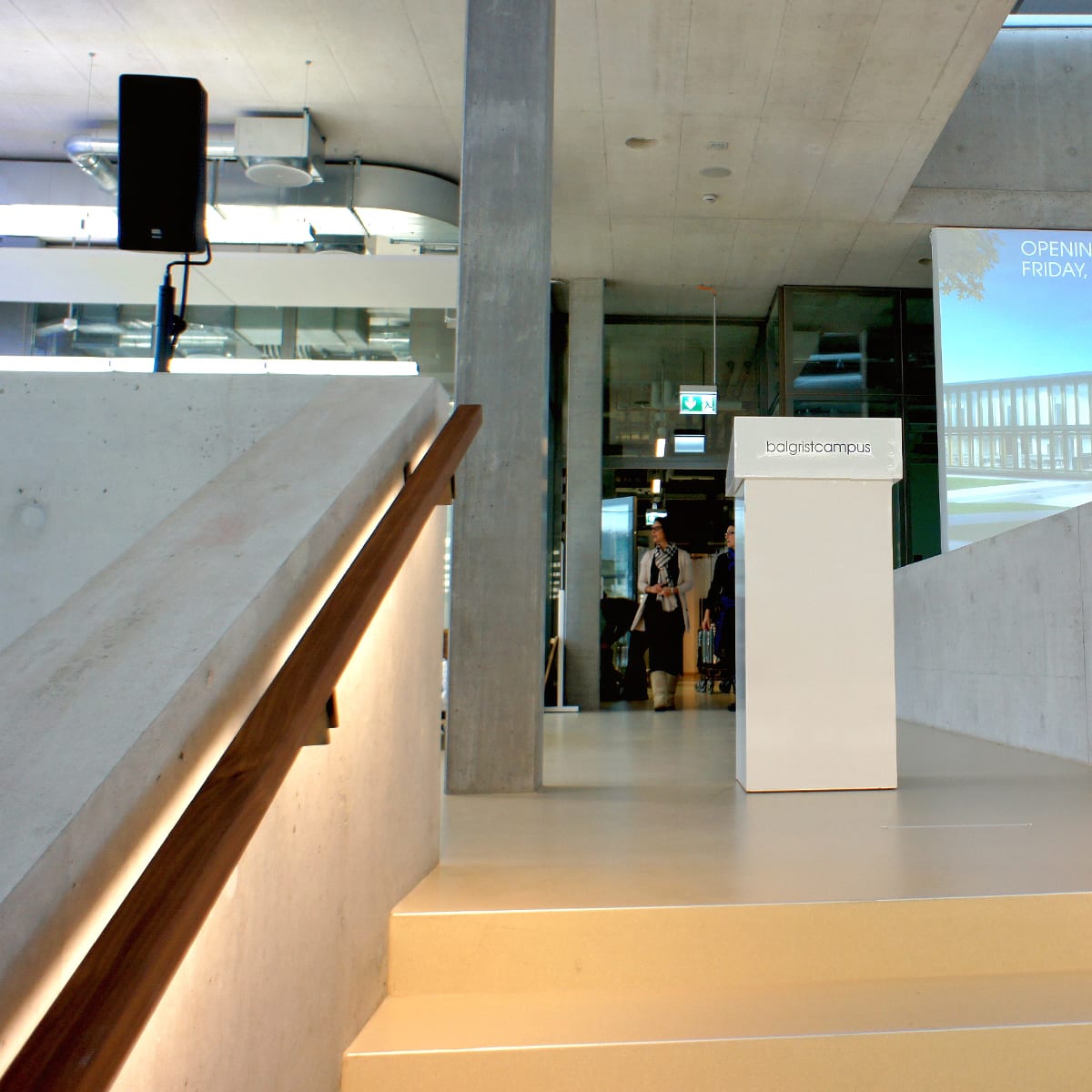 Foyer mit Betonwänden, Holzhandlauf an der Treppe und Glastüren, links oben ein Lautsprecher an der Wand